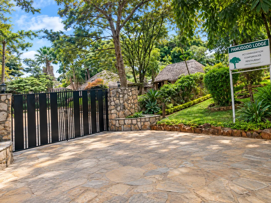 Stone-walled garden entrance with a black gate, green lawns and a Pamugodo Lodge sign reading Accommodation, Conferences, Restaurant