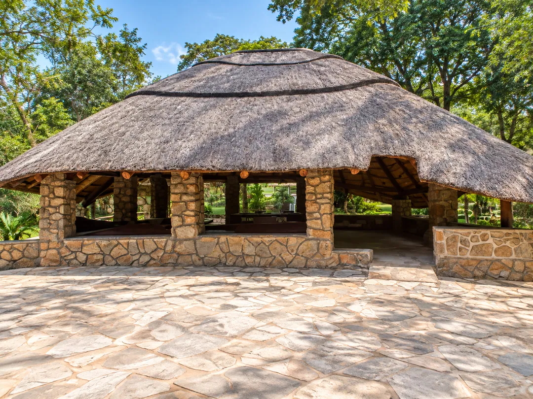 Open-sided thatched pavilion with stone pillars on a paved terrace, surrounded by leafy gardens